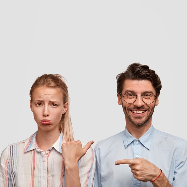 Tiro vertical de mujer rubia insatisfecha frunce el labio inferior, ofendido por el chico. Hipster sin afeitar positivo con sonrisa agradable, apunta a la novia, aislado sobre una pared blanca, espacio libre