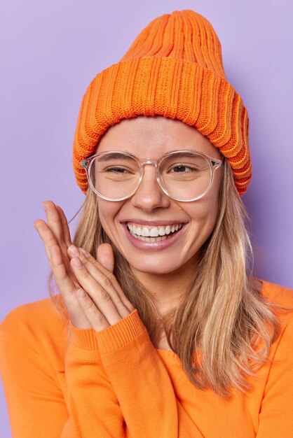 Tiro vertical de una mujer sincera y feliz que mantiene las manos juntas sonríe en términos generales recuerda algo agradable tiene un estado de ánimo optimista usa gafas ópticas grandes, sombrero naranja y un jersey de manga larga aislado sobre una pared púrpura