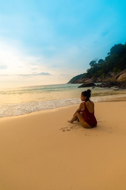 Tiro vertical de una mujer vistiendo un traje de baño rojo sentado en la playa de arena durante el amanecer