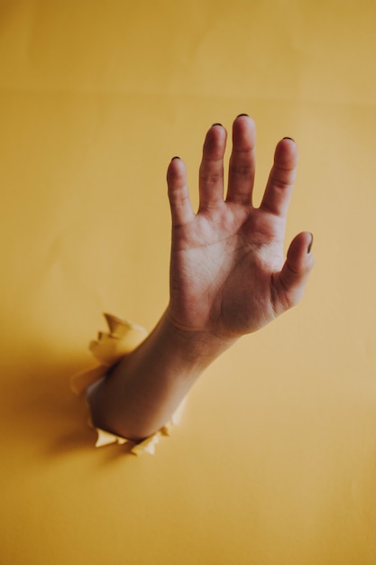 Foto gratuita tiro vertical de la palma de la mano de una persona rompiendo una pared de papel amarillo