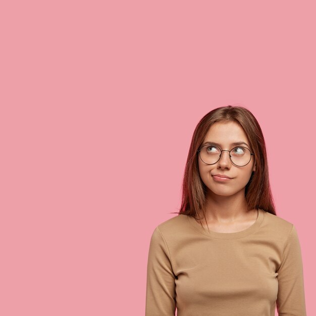 Tiro vertical de pensativa joven hermosa concentrada hacia arriba, mira dudosa y pensativa, vestida con ropa casual, tiene gafas redondas, aisladas sobre una pared rosa.