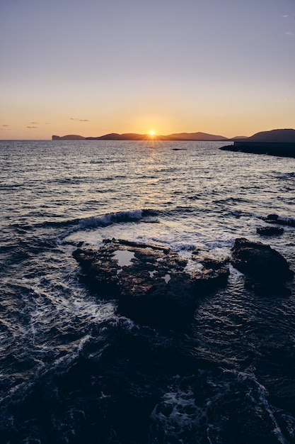 Tiro vertical de rocas en el mar con el sol brillando detrás de las montañas en la distancia