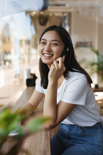 Tiro vertical sonriente, milenaria linda chica asiática en café, hablando por teléfono inteligente.