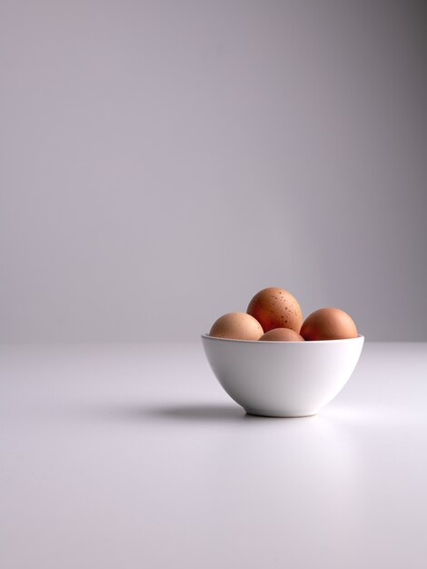 Tiro vertical de un tazón blanco con huevos marrones en una superficie blanca y fondo gris limpio