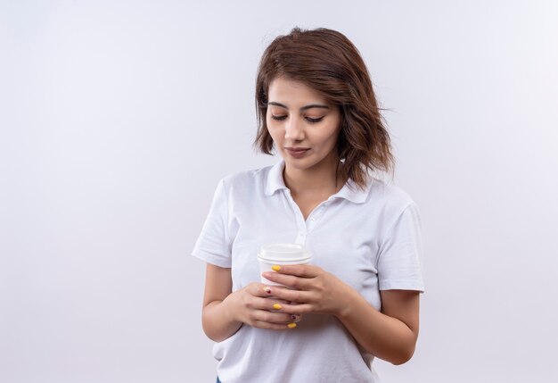 Titmid joven con pelo corto vistiendo polo blanco sosteniendo la taza de café mirando hacia abajo