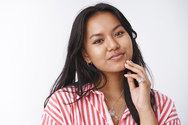 Tnder hermosa y femenina mujer asiática en blusa a rayas inclinando la cabeza suavemente tocando la cara antes de ponerse la máscara facial anti acné sonriendo encantadora y mirando a la cámara sobre una pared blanca