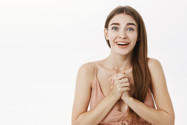 Tocada y encantada agradecida joven feliz en vestido beige apretando las manos cerca del pecho y sonriendo agradecida de ser encantado con gran sorpresa sobre la pared gris