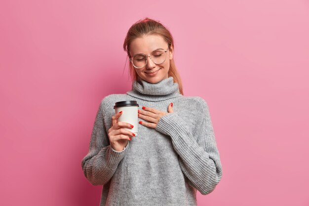 Tocada mujer europea presiona la mano contra el pecho, sostiene café para llevar, vestida con un jersey de gran tamaño, recuerda algo agradable