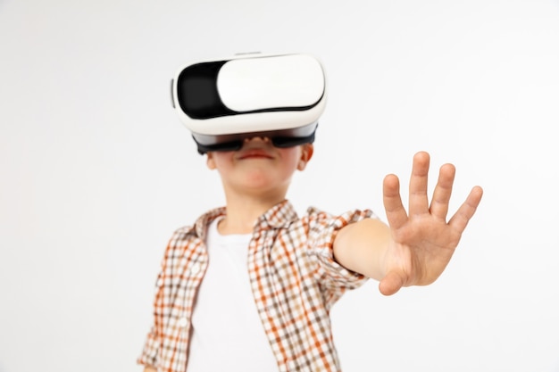 Tocando el milagro. Niño o niño en jeans y camisa con gafas de casco de realidad virtual aisladas sobre fondo blanco de estudio. Concepto de tecnología de punta, videojuegos, innovación.