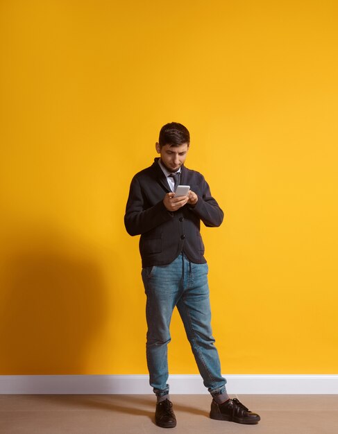 Toda la vida en gadget. Hombre caucásico joven con smartphone, sirviendo, charlando, apostando. Retrato de cuerpo entero aislado en la pared amarilla. Concepto de tecnologías modernas, millennials, redes sociales.