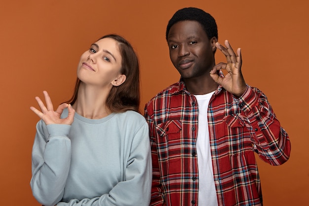 Foto gratuita todo esta bien. retrato de la feliz pareja interracial joven de buen humor dando vueltas a los pulgares con los dedos índices y sonriendo, mostrando un gesto aceptable, llenos de alegría con comprensión y apoyo mutuos