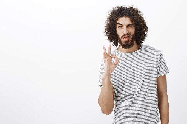 Todo bajo control, cariño. Apuesto hombre hispano confiado con barba y peinado rizado, mostrando el signo de ok o bien, guiñando un ojo