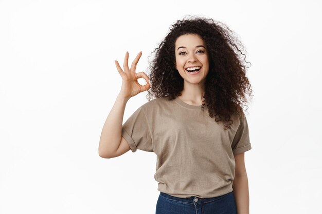 Todo bajo control. Una joven sonriente asegura que todo está bien, muestra un signo correcto y dice sí, aprueba o está de acuerdo, recomienda algo bueno, elogia el buen trabajo, de pie sobre fondo blanco