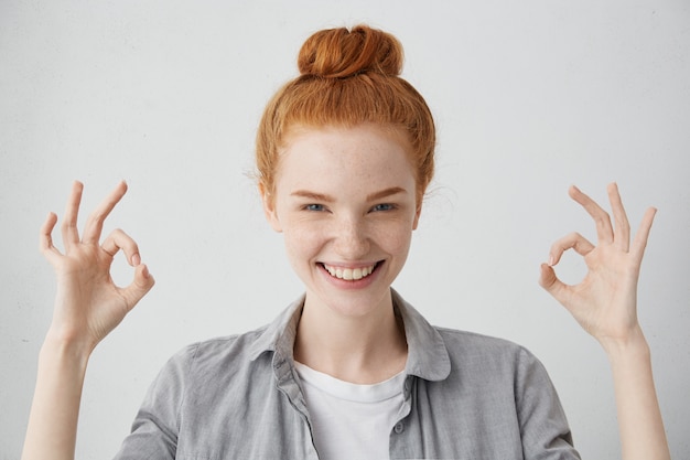 ¡Todo está bien! Alegre mujer caucásica joven emocionada con un nudo de pelo de jengibre y piel pecosa que muestra el gesto de Ok con ambas manos y una amplia sonrisa, disfrutando de su vida feliz sin preocupaciones