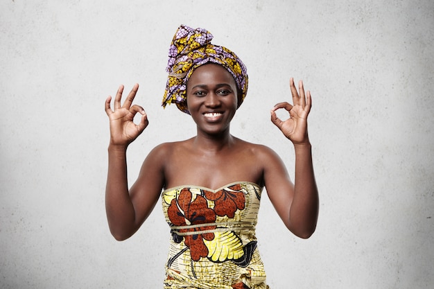 ¡Todo está bien! Hermosa mujer africana alegre con un pañuelo brillante en la cabeza y un vestido elegante que muestra el signo de ok demostrando su satisfacción y felicidad de acuerdo con algo.