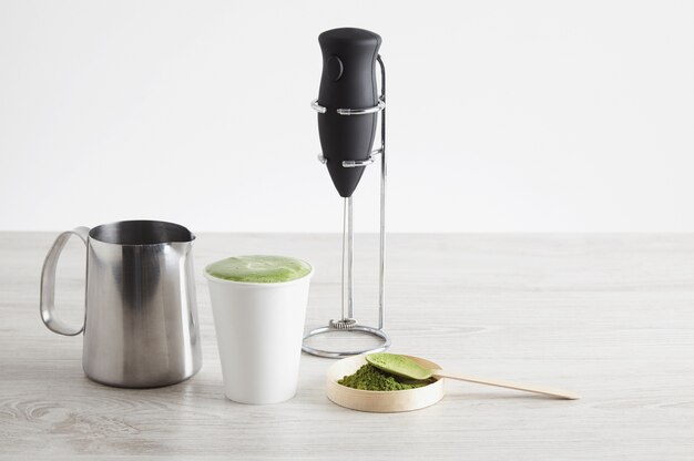 Todo lo necesario para preparar café con leche de forma moderna. Presentación de venta. Lechera eléctrica en soporte cromado, té matcha orgánico en polvo de primera calidad de Japón, vaso de papel para llevar, recipiente de acero para leche para cappuccinato