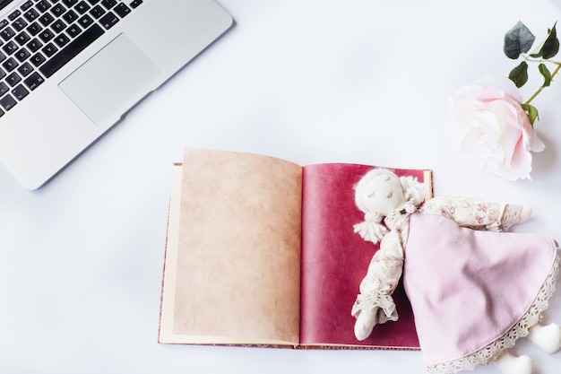 Toma aérea de un escritorio blanco con una muñeca encima de un cuaderno, una flor y una computadora portátil