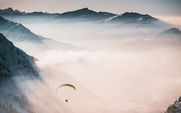 Toma aérea de una persona en paracaídas sobre las nubes cerca de montañas nevadas