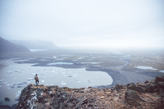Toma aérea de una persona de pie sobre un acantilado con vistas a los lagos en la niebla capturada en Islandia