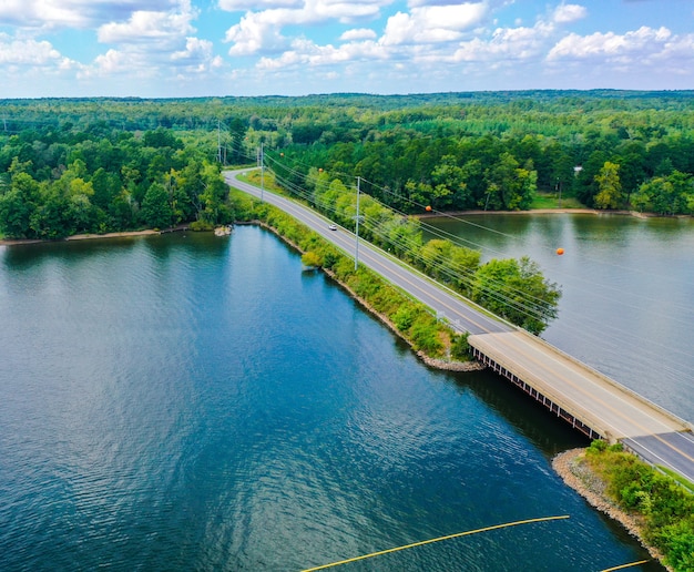 Toma aérea de un puente, camino, árboles cerca del lago con un cielo azul nublado