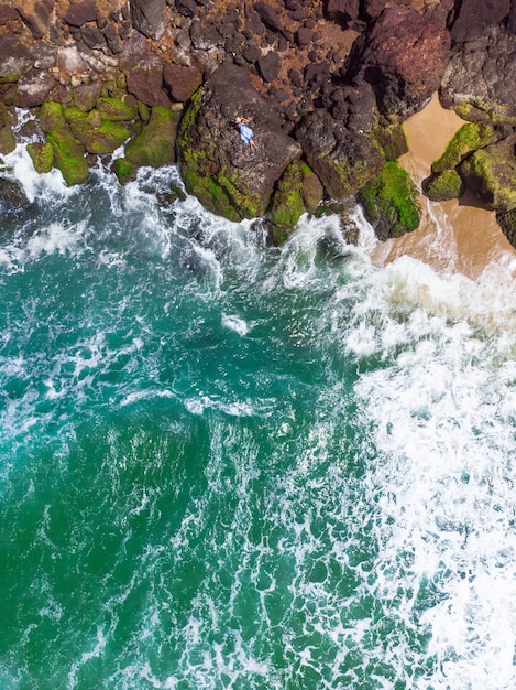 Toma aérea vertical de una mujer con vestido azul tumbado en la playa rocosa