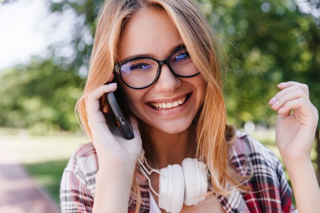 Toma al aire libre de primer plano de mujer joven soñadora en gafas hablando por teléfono. Riendo a niña extática en auriculares posando en la naturaleza borrosa.