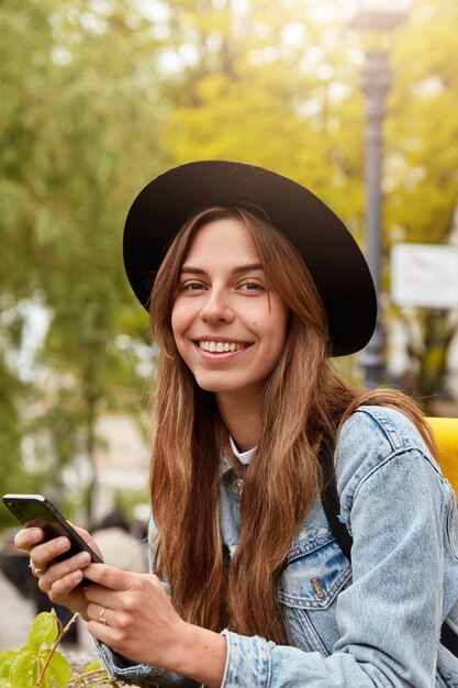 Toma exterior de feliz mujer europea con una sonrisa agradable, tiene un teléfono celular moderno, verifica la casilla de correo electrónico, disfruta de un día soleado, mensaje de texto