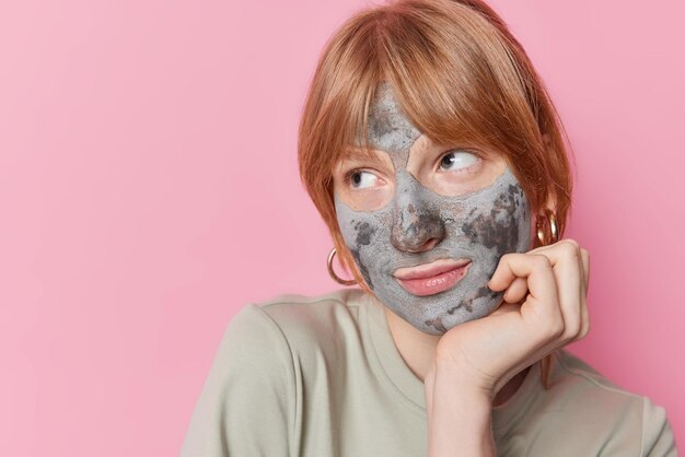 La toma horizontal de una encantadora y pensativa chica jengibre mantiene la mano bajo la barbilla enfocada y aplica una máscara de arcilla de belleza para reducir las espinillas y los poros vestidos casualmente aislados sobre un espacio de copia de fondo rosa