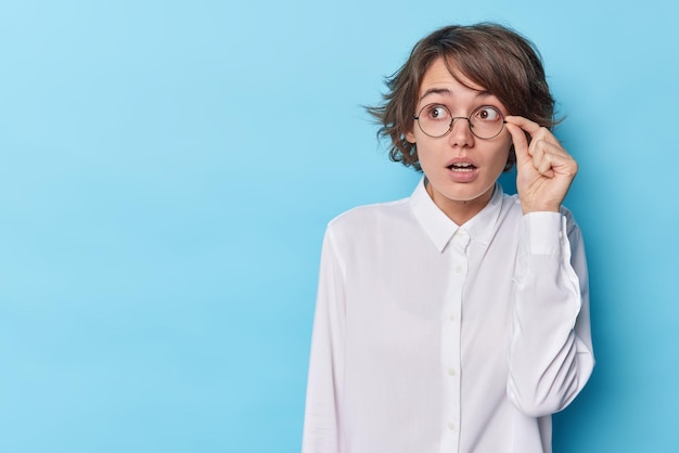 La toma horizontal de una mujer aturdida se queda sin palabras, sostiene el aliento, mantiene la mano en el borde de las gafas, usa una camisa blanca enfocada lejos, aislada sobre un fondo azul, un espacio vacío en blanco para su promoción.