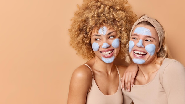 Toma horizontal de mujeres jóvenes positivas que aplican máscaras de belleza nutritivas para el cuidado de la cara y se ven con gusto para someterse a tratamientos de piel aislados sobre el espacio de copia de fondo marrón para su contenido publicitario