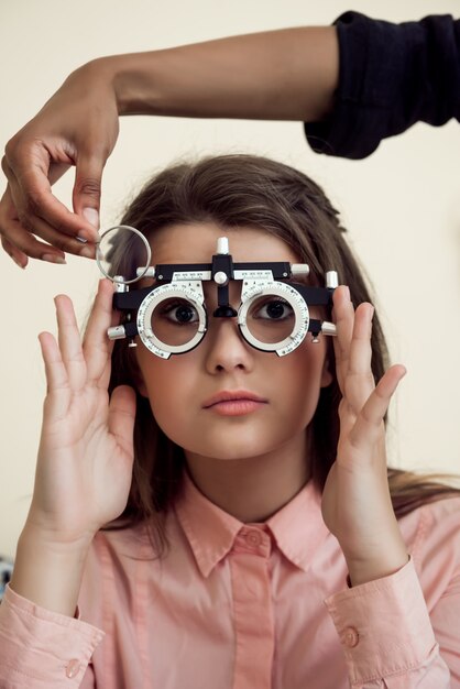 Toma horizontal de una niña caucásica interesada y curiosa en una cita con un especialista en cuidado de los ojos usando foróptero mientras el oftalmólogo revisa su visión, sentada sobre una pared amarilla