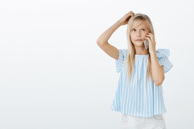 Toma interior de una adorable niña interrogada confundida con cabello rubio en una blusa azul, rascándose la cabeza y mirando hacia arriba mientras habla por teléfono inteligente, pensando lo que quiere pedirle a la abuela