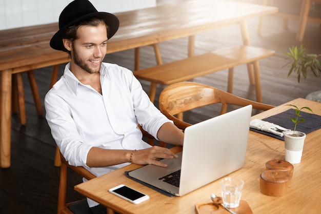Toma interior de un blogger masculino escribiendo en el teclado de la computadora portátil, usando wi-fi gratis en un café moderno mientras trabaja en su nueva publicación en las redes sociales, mirando la pantalla con una expresión de rostro feliz e inspirada