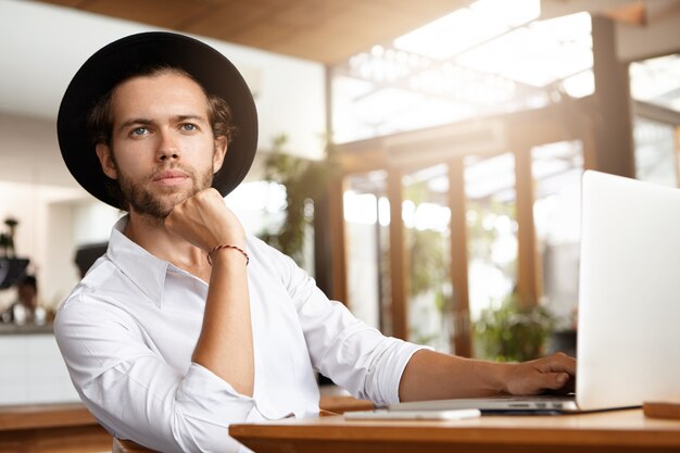 Toma interior de un bloguero joven y guapo con sombreros trabajando en una nueva publicación para su blog usando wi-fi en una computadora portátil genérica, sosteniendo la mano en su barbilla y mirando hacia adelante con expresión pensativa