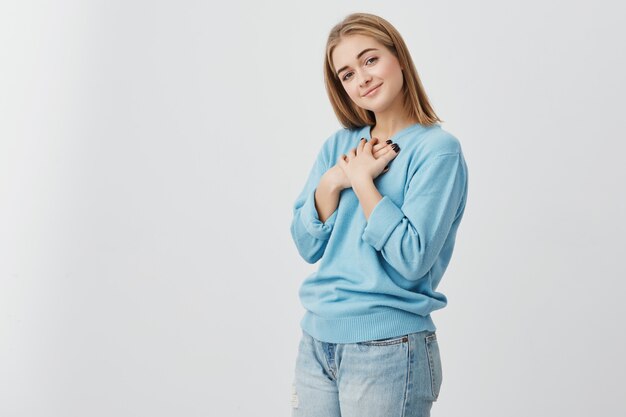 Toma interior de una chica estudiante muy hermosa con el pelo rubio y liso sonriendo alegremente, manteniendo las manos sobre su pecho, satisfecha con un regalo inesperado. Expresiones y emociones humanas positivas.