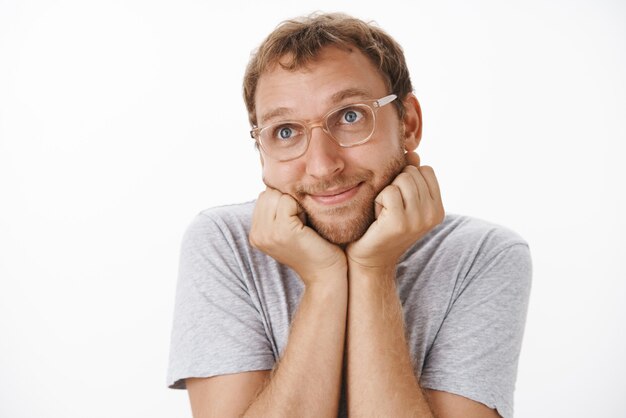 Toma interior de un chico adulto lindo y tierno con gafas y camiseta gris sonriendo con deleite, apoyando la cabeza en las palmas y mirando la esquina superior izquierda nostálgica y romántica