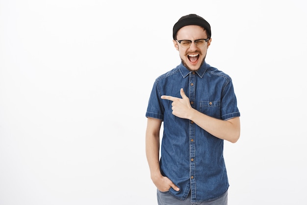 Toma interior de un chico barbudo hipster guapo emocionado y sorprendido con gorro negro gritando en voz alta con los ojos cerrados y la boca abierta apuntando hacia la izquierda abrumado por las emociones sobre la pared blanca