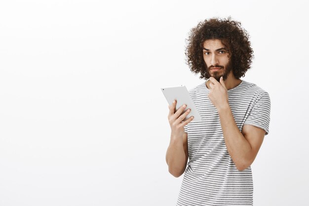 Toma interior de un chico guapo serio pensativo con cabello rizado, tocando la barba y mirando enfocado mientras piensa, sosteniendo una tableta digital blanca