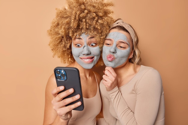 Toma interior de dos modelos femeninas jóvenes que toman una pose selfie en un teléfono inteligente en la cámara frontal y aplican una máscara nutritiva de belleza en las caras vestidas casualmente aisladas sobre un fondo marrón Concepto de cuidado de la piel