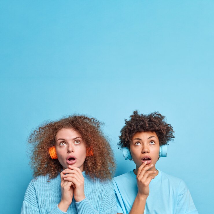 Foto gratuito toma interior de dos mujeres jóvenes de raza mixta concentradas en lo alto con expresiones de asombro y asombro escuchar música en auriculares inalámbricos notar algo asombroso aislado sobre una pared azul