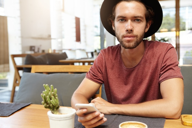 Toma interior de un estudiante elegante con sombrero negro enviando mensajes de texto a sus amigos a través de las redes sociales, usando wi-fi gratis en su teléfono celular durante el desayuno en la cafetería con un interior moderno, mirando