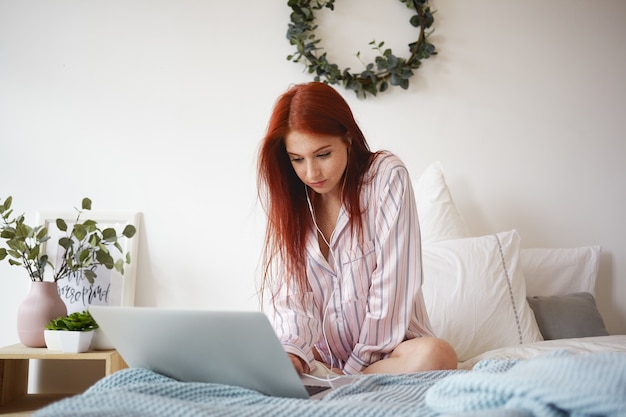 Toma interior de una estudiante pelirroja trabajadora en ropa de dormir elegante sentada en la cama deshecha con auriculares, usando una conexión inalámbrica a Internet de alta velocidad en una computadora portátil mientras realiza tareas en el hogar