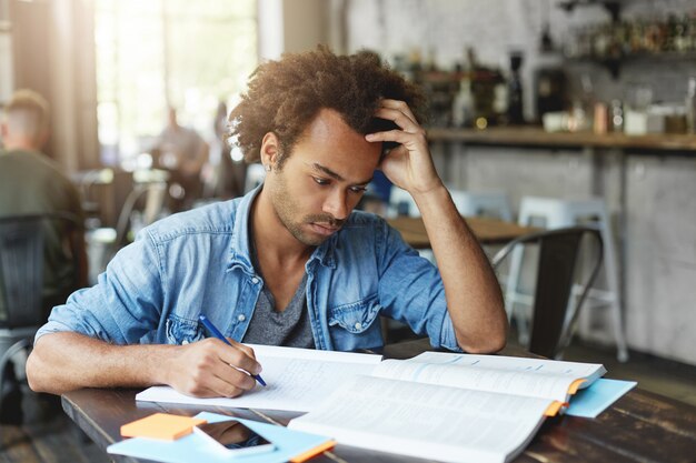 Toma interior de un estudiante universitario europeo negro elegante y guapo enfocado que tiene una expresión facial concentrada seria mientras aprende una lección en el café por la mañana, resolviendo problemas matemáticos