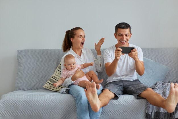 Toma interior de familia emocionada sentada en un sofá en la sala de estar, esposo sosteniendo el teléfono móvil en las manos, teniendo excelentes noticias sobre su victoria en la lotería, gente con infante gritando wow felizmente.