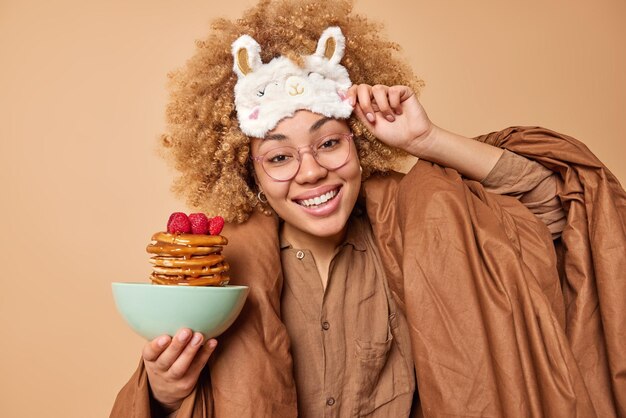 Toma interior de feliz y encantadora mujer joven despierta feliz después de dormir weas pijama posa bajo una manta que va a tomar un delicioso desayuno sostiene un tazón con panqueques aislados sobre la pared beige del estudio