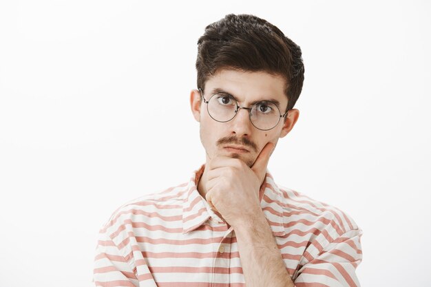 Toma interior de un hermano mayor enojado enfocado serio con bigote en anteojos de moda, sosteniendo la mano en la barbilla y mirando con expresión molesta disgustada, resolviendo un problema difícil sobre una pared gris