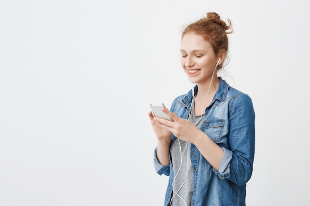 Toma interior de una hermosa adolescente pelirroja con el pelo peinado en un moño, sonriendo ampliamente mientras envía mensajes de texto o navega en la red social a través de un teléfono inteligente