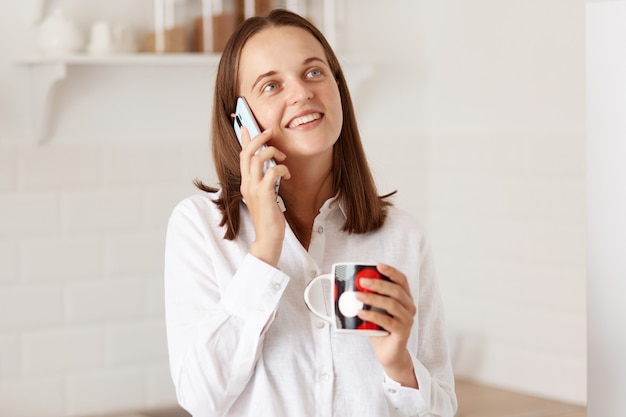 Toma interior de hermosa chica hablando por teléfono móvil, sosteniendo una taza de café o té, mirando a otro lado y sonriendo mientras posa con la cocina en segundo plano.