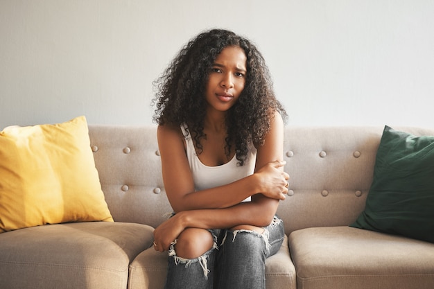 Toma interior de una hermosa joven de raza mixta de moda con peinado afro sentada en el sofá en casa, frunciendo el ceño, con mirada triste y preocupada, sufriendo de calambres estomacales o sintiéndose sola y molesta