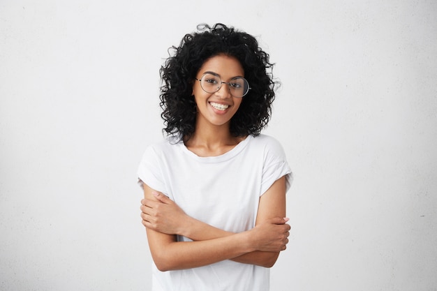 Toma interior de hermosa mujer afroamericana feliz sonriendo alegremente, manteniendo los brazos cruzados, relajándose en el interior después de las conferencias matutinas en la universidad